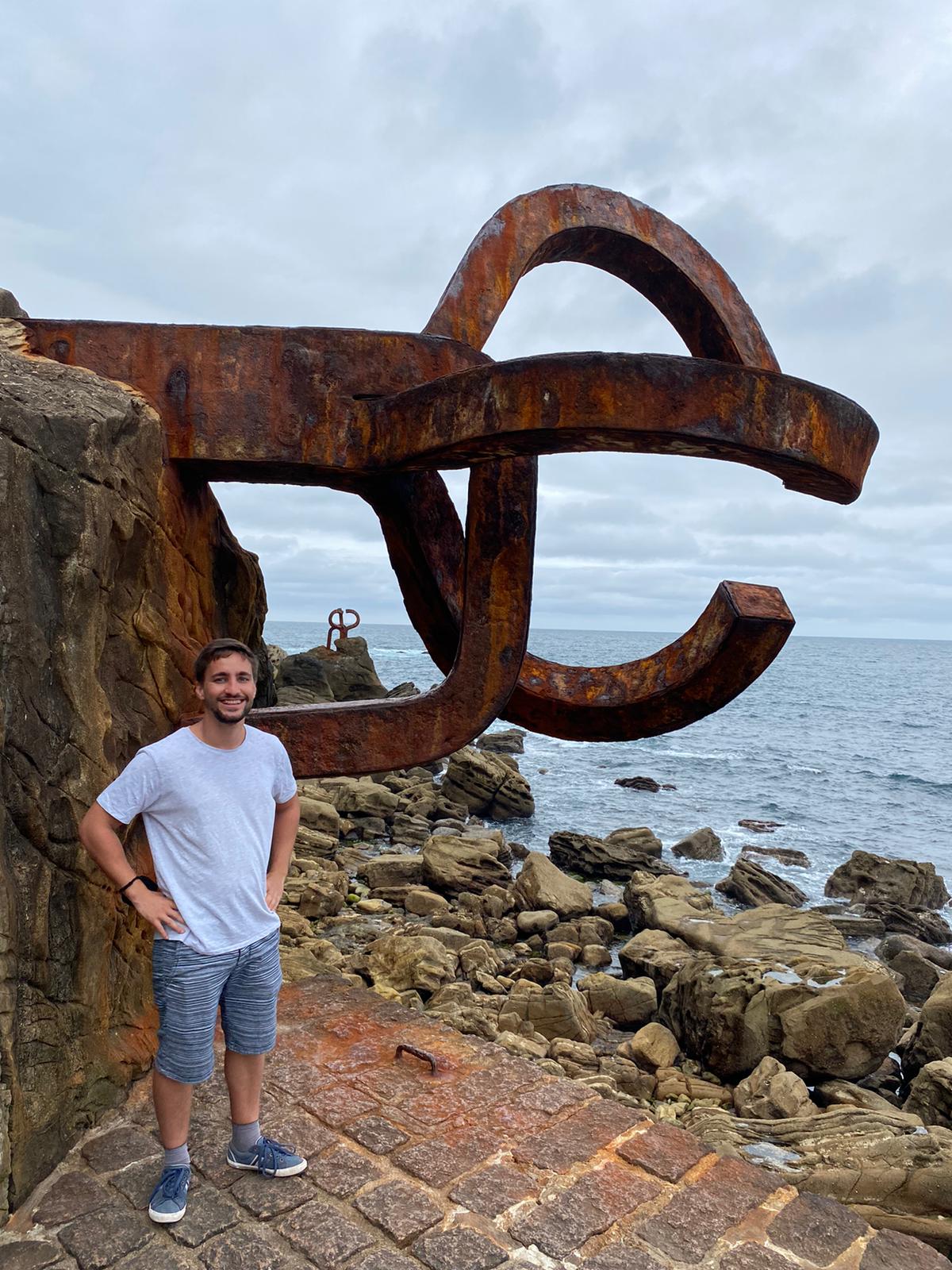 Alfonso standing beside a large abstract steel sculpture on the coast